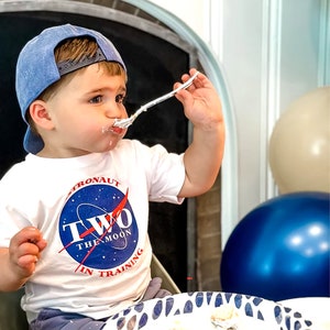 May include: A toddler wearing a white t-shirt with a blue and red astronaut logo that says "Two the Moon in Training" and a blue baseball cap is eating cake with a spoon.