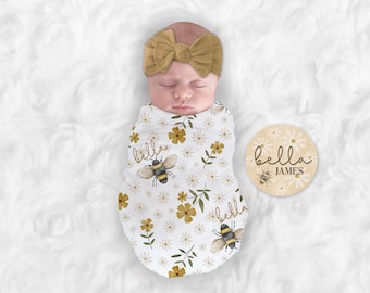 May include: A newborn baby wrapped in a white swaddle with a yellow and white floral pattern and bees. The baby is wearing a brown headband with a bow. A wooden round sign with a white floral pattern and bees says "bella JAMES".
