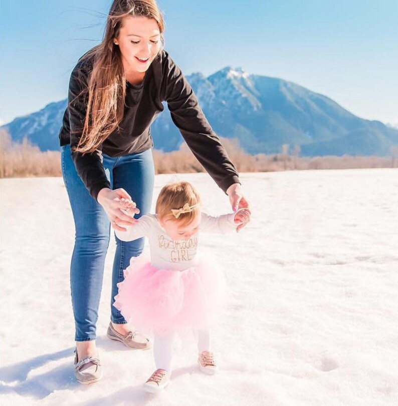 May include: A young girl in a pink tutu and a white shirt with gold lettering that says "Birthday Girl" is taking her first steps while being helped by an adult woman in a black sweater and blue jeans. The background is a snowy field with mountains in the distance.