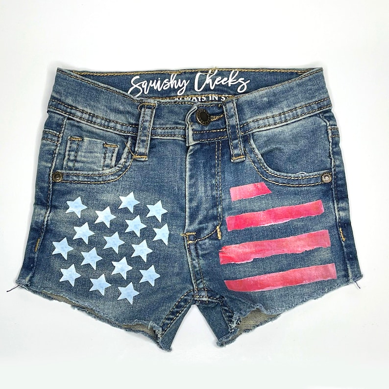 Pode incluir: Cal&ccedil;as de ganga azuis com um design de bandeira americana pintada. A bandeira &eacute; pintada em vermelho, branco e azul. As cal&ccedil;as t&ecirc;m uma bainha desfiada e s&atilde;o desgastadas.