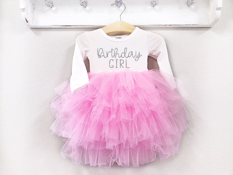 May include: A white long-sleeved shirt with a pink tulle skirt. The shirt has the text "Birthday Girl" in silver glitter.