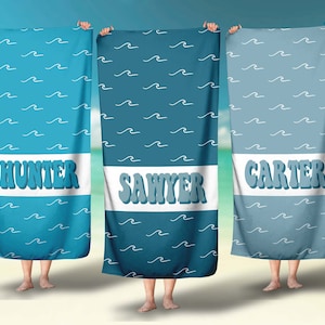 May include: Three blue beach towels with white wave patterns and personalized names: Hunter, Sawyer, and Carter. The towels are held up by hands in front of a beach background.