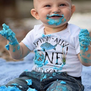 Puede incluir: Un beb&eacute; var&oacute;n con un body blanco con el texto "Got Fish-ally One" y pantalones vaqueros azules est&aacute; cubierto de glaseado azul. El beb&eacute; sonr&iacute;e y tiene glaseado en la cara y las manos.