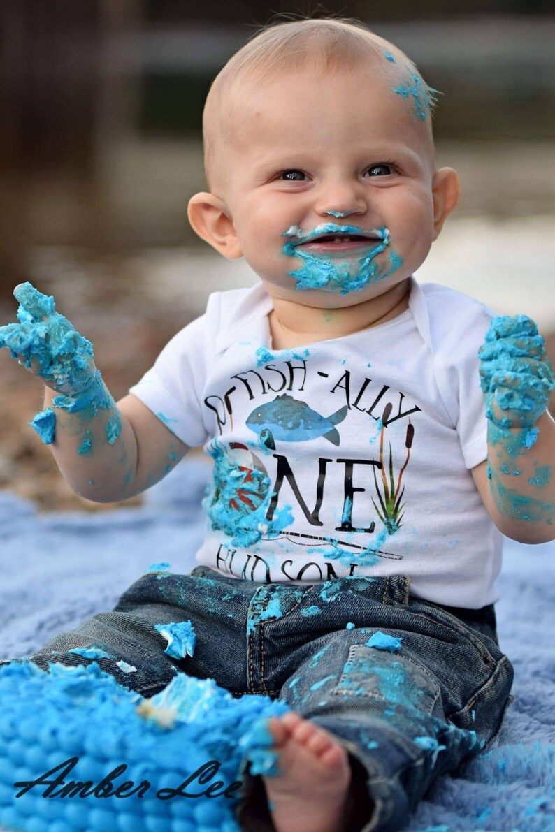 Puede incluir: Un beb&eacute; var&oacute;n con un body blanco con el texto "Got Fish-ally One" y pantalones vaqueros azules est&aacute; cubierto de glaseado azul. El beb&eacute; sonr&iacute;e y tiene glaseado en la cara y las manos.