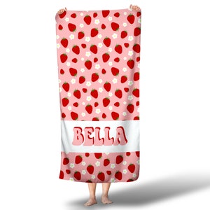 May include: Pink beach towel with a white border and a pattern of red strawberries and white flowers. The name "BELLA" is printed in a retro font on the white border.
