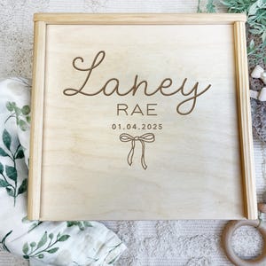 Puede incluir: Una caja de madera para recuerdos con una inscripción personalizada. La inscripción dice "Laney Rae 01.04.2025" con un lazo decorativo.