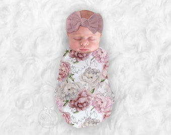 May include: A white swaddle with a floral pattern featuring pink and gray peonies. The swaddle is personalized with the name "Brinkley" in a script font. A baby is wearing the swaddle and a pink bow headband.