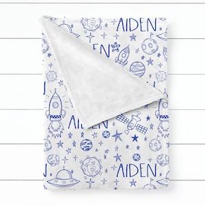Puede incluir: Una manta de forro polar blanca con un patrón azul de temática espacial. El patrón incluye cohetes, planetas, estrellas y el nombre "Aiden".