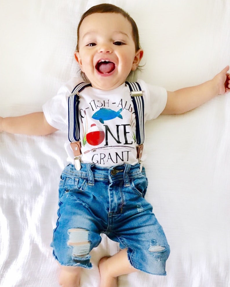 Puede incluir: Un beb&eacute; var&oacute;n con un body blanco con tirantes azules y blancos. El body tiene un gr&aacute;fico de un pez y el texto "O-FISH-ALLY ONE GRANT". El beb&eacute; lleva pantalones cortos de mezclilla azul con detalles desgastados.