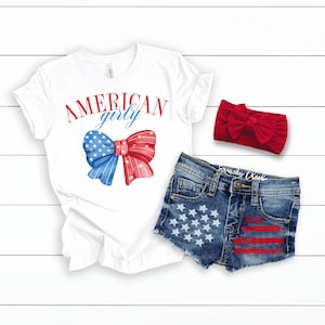 Pode incluir: Camiseta branca com o texto "AMERICAN girly" e um la&ccedil;o vermelho, branco e azul. Cal&ccedil;as de ganga azuis com estrelas brancas e riscas vermelhas. Faixa de cabe&ccedil;a vermelha.