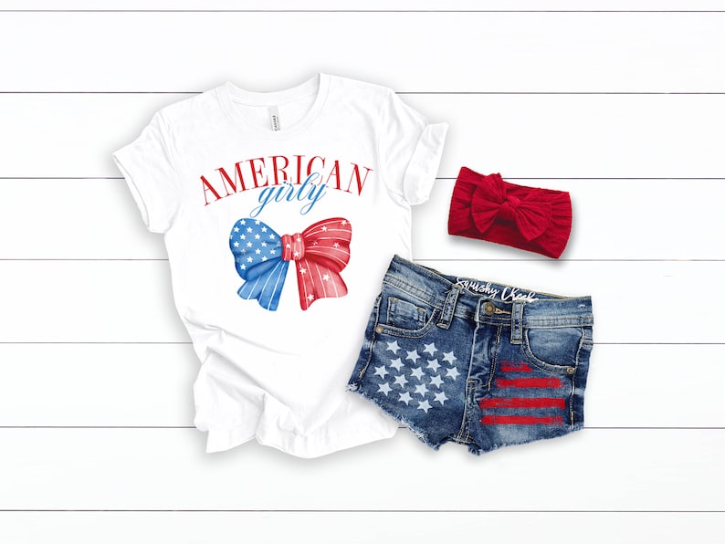 Pode incluir: Camiseta branca com o texto "AMERICAN girly" e um la&ccedil;o vermelho, branco e azul. Cal&ccedil;as de ganga azuis com estrelas brancas e riscas vermelhas. Faixa de cabe&ccedil;a vermelha.