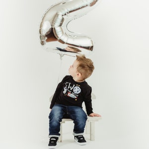 May include: A young boy wearing a black long-sleeved shirt with a graphic of an astronaut and the text "Two the Moon Nixon" sits on a white stool. A silver number two balloon is above his head.