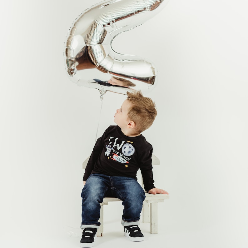 May include: A young boy wearing a black long-sleeved shirt with a graphic of an astronaut and the text "Two the Moon Nixon" sits on a white stool. A silver number two balloon is above his head.