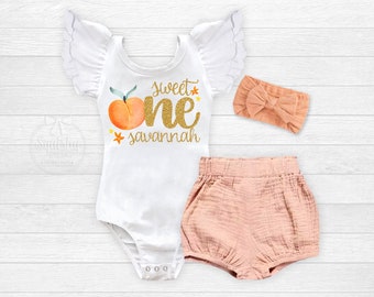 Op de afbeelding: Witte romper met gouden glittertekst die "Sweet One Savannah" zegt en een perzikgrafiek. Een perzikkleurig hoofdband en bijpassende shorts zijn ook inbegrepen.