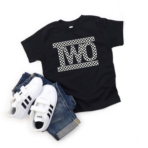 May include: Black t-shirt with a white "TWO" graphic in a checkered pattern. The shirt is on a white background with a pair of white sneakers and blue jeans.