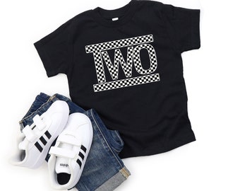 Puede incluir: Camiseta negra con un gráfico blanco "DOS" en un patrón de cuadros. La camiseta está sobre un fondo blanco con un par de zapatillas blancas y pantalones vaqueros azules.