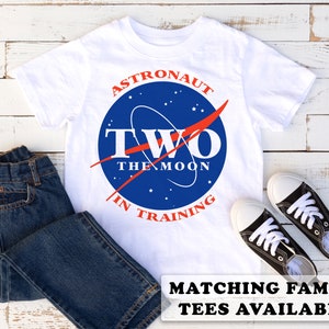 May include: White t-shirt featuring a blue and red astronaut logo that reads "Astronaut Two The Moon In Training". The shirt is displayed on a white wooden background with blue jeans and black trainers.