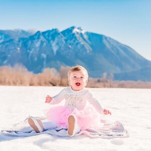 May include: A baby girl wearing a white long-sleeved shirt with the text "1st Birthday Girl" in gold glitter, a pink tulle tutu, and white shoes, sitting on a white blanket in the snow with a mountain range in the background.