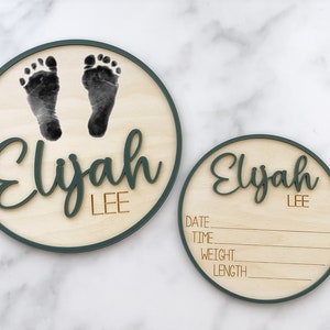 May include: Two round wooden plaques with a green border. The larger plaque has a pair of baby footprints and the name "Elijah LEE" in green lettering. The smaller plaque has the name "Elijah LEE" and blank lines for date, time, weight, and length.