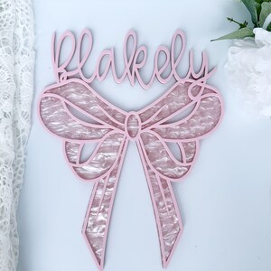 May include: Pink wooden sign with the name "Blakely" written in cursive script. The sign features a large pink bow with a pearl-like texture.