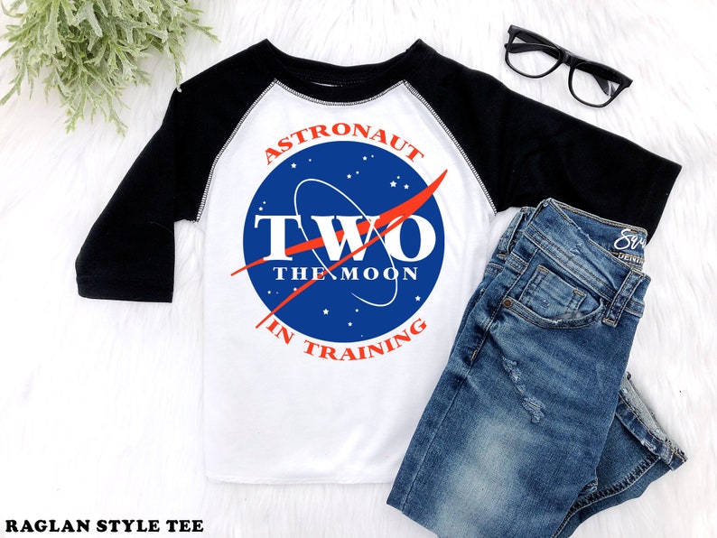 May include: A white and black raglan style tee shirt with a blue and red NASA-inspired logo that says "Astronaut Two The Moon In Training".