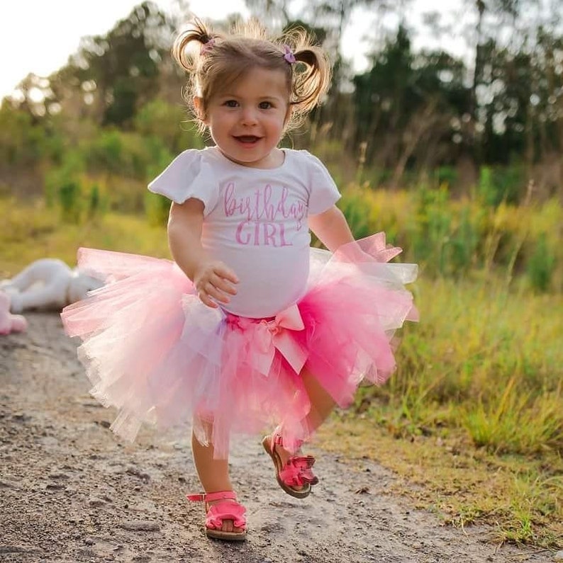 May include: A young girl wearing a white shirt with the text "birthday girl" in pink lettering, a pink and white tulle tutu skirt, and pink shoes. She is standing on a dirt path with green grass in the background.