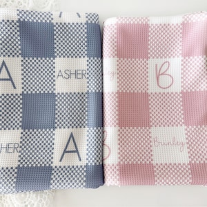 Puede incluir: Dos mantas de bebé personalizadas con un patrón a cuadros. Una manta es azul con el nombre "Asher" y la otra es rosa con el nombre "Brinkley".