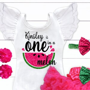 May include: A white onesie with a pink glitter watermelon graphic and the text "Kinsley is one in a melon". The onesie has white ruffled sleeves and is paired with a pink tulle skirt and a headband with a pink glitter bow. There are also two pink flower hair clips with green leaves.