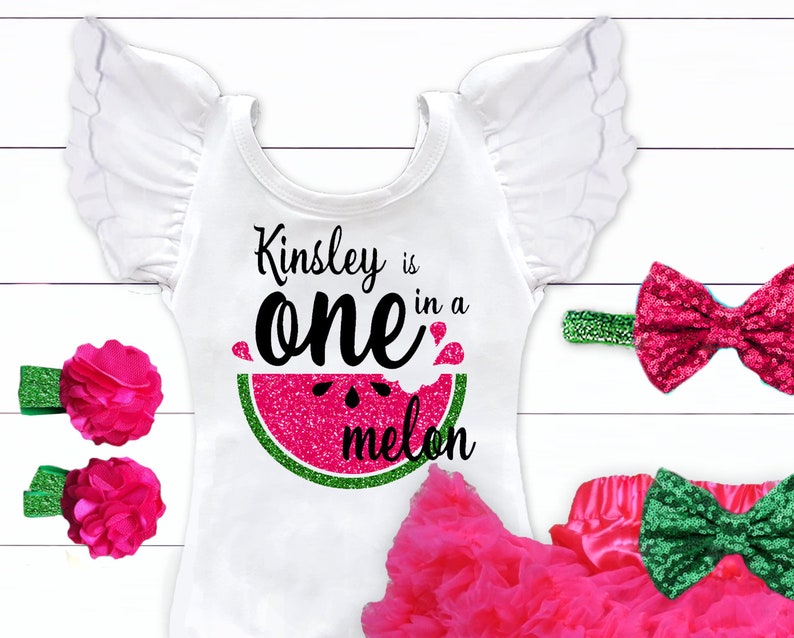 May include: A white onesie with a pink glitter watermelon graphic and the text "Kinsley is one in a melon". The onesie has white ruffled sleeves and is paired with a pink tulle skirt and a headband with a pink glitter bow. There are also two pink flower hair clips with green leaves.