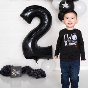 May include: A young child wearing a black long-sleeved shirt with a graphic of an astronaut on the moon and the text "Two the Moon Ethan". The child is standing in front of a large black number two balloon and a white balloon.