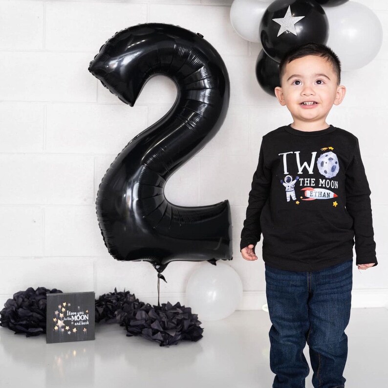 May include: A young child wearing a black long-sleeved shirt with a graphic of an astronaut on the moon and the text "Two the Moon Ethan". The child is standing in front of a large black number two balloon and a white balloon.