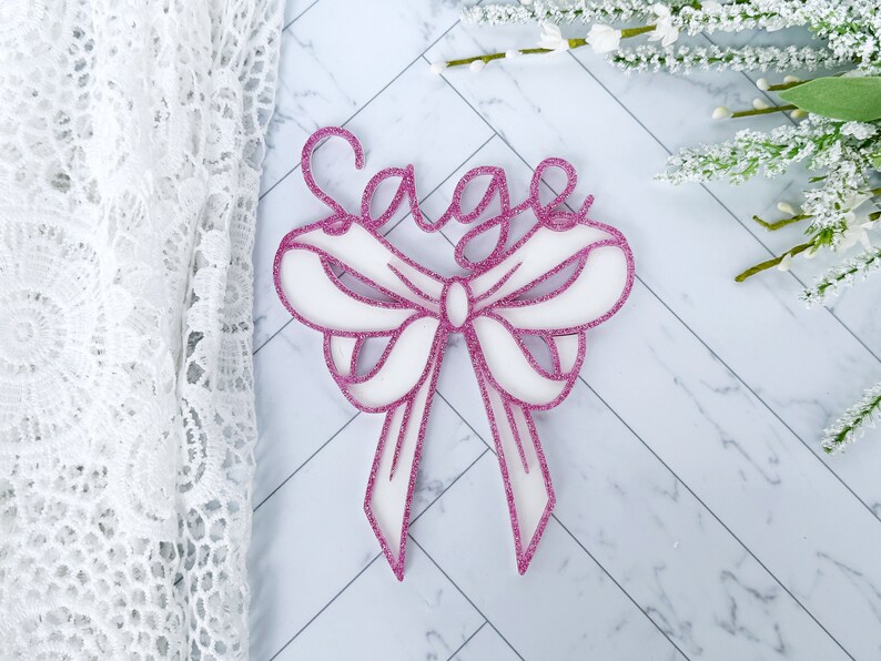 May include: A pink glitter bow with the name "Sage" written in white glitter on top.
