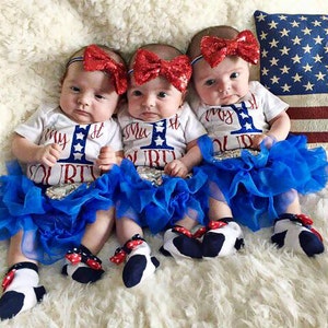 Puede incluir: Tres beb&eacute;s con monos blancos con detalles en rojo, blanco y azul y el texto "My 1st Fourth" en ellos. Llevan tut&uacute;s de tul azul y diademas rojas, blancas y azules con lazos. Los beb&eacute;s est&aacute;n sentados sobre una superficie blanca y esponjosa.