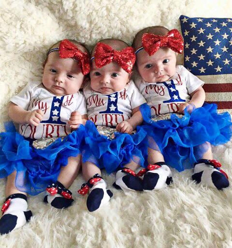 Puede incluir: Tres beb&eacute;s con monos blancos con detalles en rojo, blanco y azul y el texto "My 1st Fourth" en ellos. Llevan tut&uacute;s de tul azul y diademas rojas, blancas y azules con lazos. Los beb&eacute;s est&aacute;n sentados sobre una superficie blanca y esponjosa.