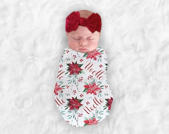Könnte beinhalten: Eine weiße Wickeldecke mit einem Muster aus roten Weihnachtssternen und grünem Laub. Die Decke trägt den Namen "Noelle". Ein Baby schläft in der Wickeldecke und trägt ein rotes Schleifenband.