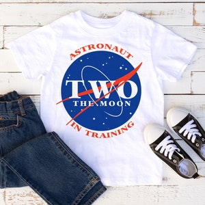 May include: White t-shirt featuring a blue and red NASA-inspired logo that reads "Astronaut Two The Moon In Training". The shirt is styled with blue jeans and black trainers.