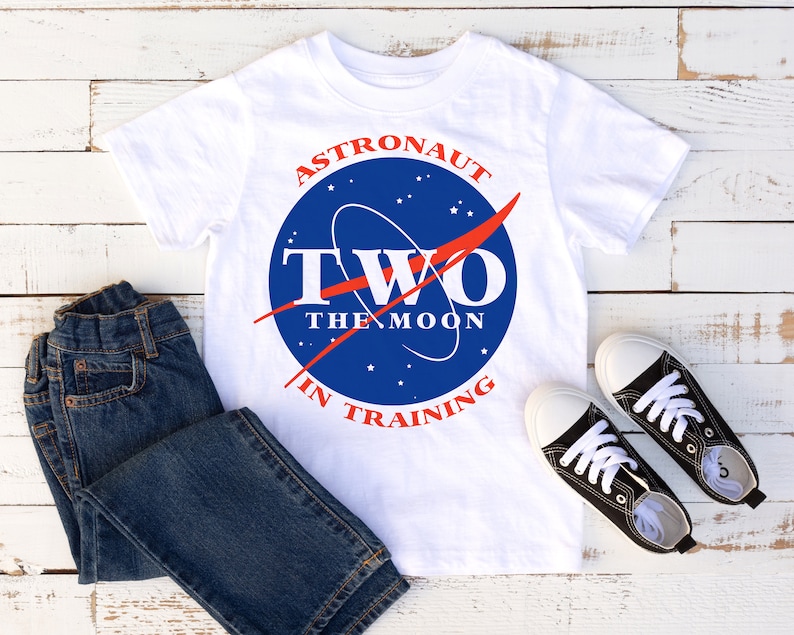 May include: White t-shirt featuring a blue and red NASA-inspired logo that reads "Astronaut Two The Moon In Training". The shirt is styled with blue jeans and black trainers.