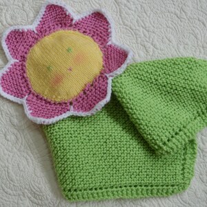 Könnte beinhalten: Eine gestrickte Babydecke mit einer rosa Blumenapplikation. Die Blume hat eine gelbe Mitte mit einem lächelnden Gesicht.