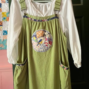 May include: A sage green pinafore dress featuring adjustable metal buckles, two side pockets, and a central circular patch with a blue bird and floral patterns. The dress is layered over a white long-sleeved blouse with a gathered neckline.