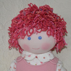 Puede incluir: Una muñeca hecha a mano con pelo de lana rosa, ojos azules y un vestido rosa y blanco.