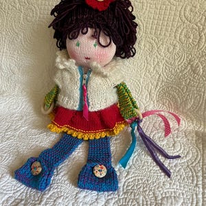 Pode incluir: Boneca feita à mão com rosto de malha, cabelo de fio roxo escuro e um acessório de cabelo de flor vermelha. A boneca usa um colete de malha branco, uma saia vermelha com acabamento amarelo e calças azuis com botões decorativos. Fitas de várias cores estão presas.