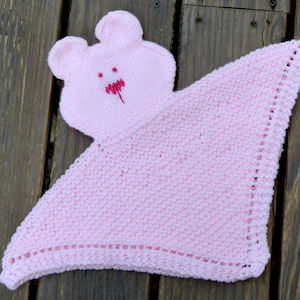 Könnte beinhalten: Eine rosa gestrickte Babydecke mit einem angestrickten rosa Bärenkopf. Der Bär hat eine rote gestickte Nase und einen Mund.