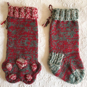 Könnte beinhalten: Zwei rot-grüne gestrickte Weihnachtsstrümpfe mit weißen Pfotenabdrücken am Boden. Die Strümpfe haben einen weißen Bund mit einem weißen Quasten.