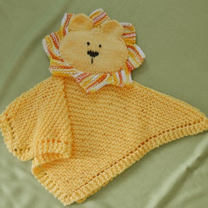 May include: A handmade, knitted baby security blanket in the shape of a lion. The lion's face is light yellow with a black nose and eyes. The mane is striped with orange, yellow, and white. The blanket is a solid light yellow color.