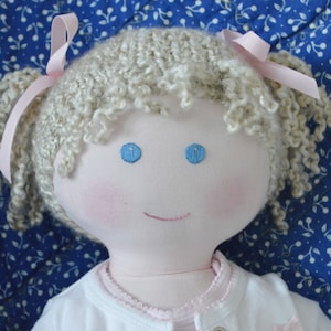 May include: A handmade doll with blonde curly hair, blue eyes, and a pink and white outfit. The doll is smiling and has rosy cheeks.