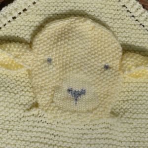 May include: A knitted baby blanket featuring a yellow lamb applique. The lamb has a grey nose and eyes.