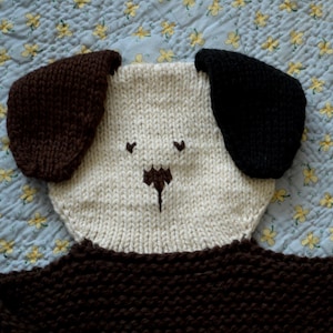 May include: A knitted dog toy with brown and black ears and a white face. The toy is made of soft yarn and has a cute, friendly expression.