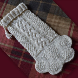 May include: A white knitted Christmas stocking with a cable knit pattern. The stocking has a loop at the top and a tassel.