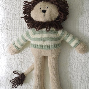 Puede incluir: Un juguete de león de punto con una melena marrón, que lleva un suéter a rayas verdes y blancas. El juguete tiene una cola larga y está de pie sobre una superficie blanca.