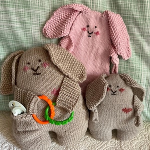 Könnte beinhalten: Drei gestrickte Spielzeug-Hasen in Rosa- und Beigetönen. Die Hasen haben gestickte Gesichter und lange, schlaffe Ohren. Ein Hase hält einen Schnuller und einen Beißring. Die Spielzeuge sind weich und kuschelig, für ein Baby oder ein Kleinkind.